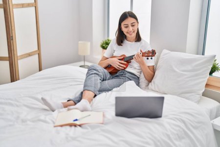 Foto de Joven mujer hispana jugando ukelele sentado en la cama en el dormitorio - Imagen libre de derechos