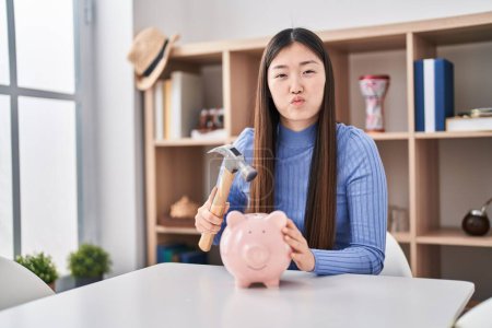 Photo for Chinese young woman holding hammer and piggy bank puffing cheeks with funny face. mouth inflated with air, catching air. - Royalty Free Image