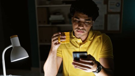 Foto de Trabajador de negocios joven latino usando teléfono inteligente bebiendo café en la oficina - Imagen libre de derechos
