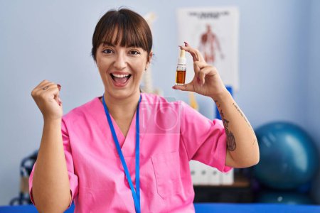 Photo for Young brunette woman holding cbd oil at physiotherapy clinic screaming proud, celebrating victory and success very excited with raised arms - Royalty Free Image