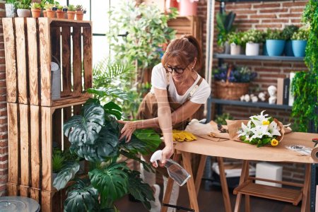 Téléchargez les photos : Femme d'âge moyen fleuriste en utilisant difusser travaillant à la boutique de fleurs - en image libre de droit