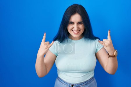 Photo for Young modern girl with blue hair standing over blue background shouting with crazy expression doing rock symbol with hands up. music star. heavy music concept. - Royalty Free Image