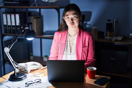 Téléchargez les photos : Jeune femme chinoise travaillant au bureau la nuit gonflant les joues avec un drôle de visage. bouche gonflée d'air, expression folle. - en image libre de droit