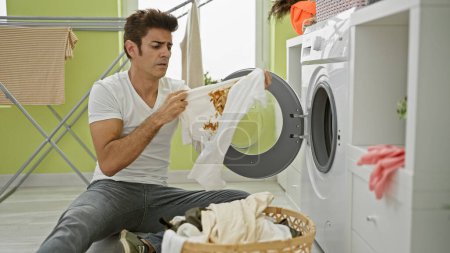 Téléchargez les photos : Jeune homme hispanique laver les vêtements tenant t-shirt sale avec tache à la buanderie - en image libre de droit