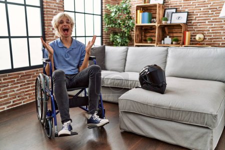 Photo for Young man sitting on wheelchair at for motorcycle accident celebrating victory with happy smile and winner expression with raised hands - Royalty Free Image