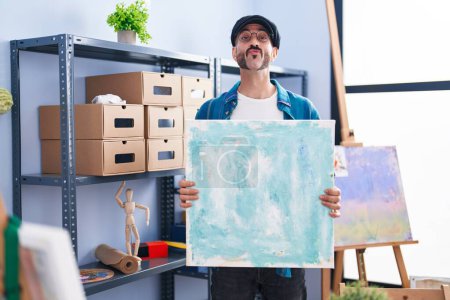 Foto de Hombre hispano con barba sosteniendo lienzo en el estudio mirando a la cámara soplando un beso siendo encantador y sexy. expresión de amor. - Imagen libre de derechos