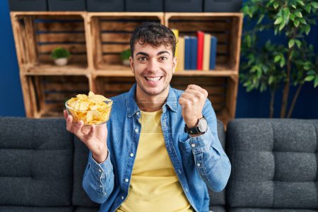 Photo for Young hispanic man holding potato chips screaming proud, celebrating victory and success very excited with raised arms - Royalty Free Image