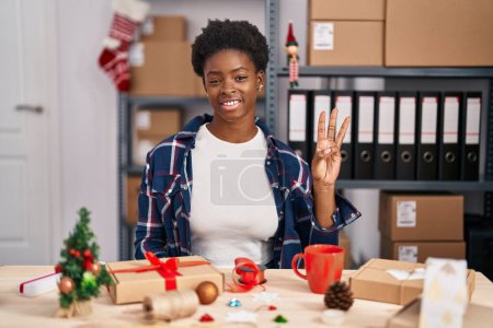 Foto de Mujer afroamericana trabajando en pequeñas empresas haciendo decoración navideña mostrando y señalando con los dedos el número tres mientras sonríe confiada y feliz. - Imagen libre de derechos