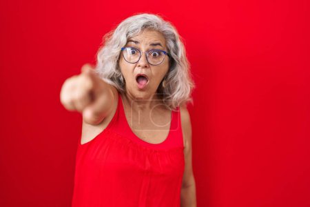 Foto de Mujer de mediana edad con el pelo gris de pie sobre fondo rojo apuntando con el dedo sorprendido por delante, boca abierta expresión asombrada, algo en la parte delantera - Imagen libre de derechos