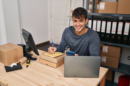 Téléchargez les photos : Jeune homme hispanique ecommerce travailleur d'affaires en utilisant l'écriture smartphone sur le paquet au bureau - en image libre de droit