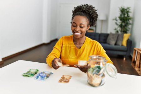 Photo for African american woman sitting on table pulling apart rand banknotes at home - Royalty Free Image
