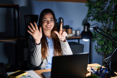 Photo for Young brunette woman working at the office at night showing and pointing up with fingers number seven while smiling confident and happy. - Royalty Free Image