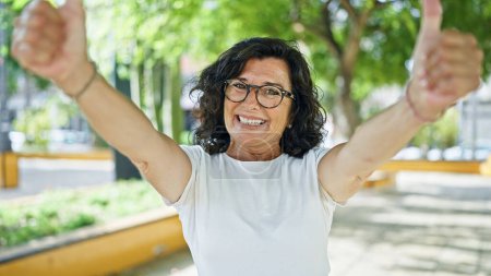 Foto de Mujer hispana de mediana edad sonriendo confiada haciendo buen signo con pulgares en el parque - Imagen libre de derechos