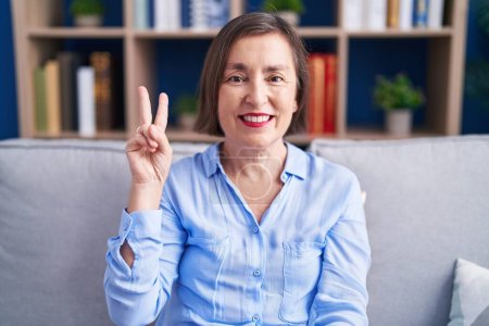 Foto de Mujer hispana de mediana edad sentada en el sofá en casa mostrando y señalando con los dedos el número dos mientras sonríe confiada y feliz. - Imagen libre de derechos