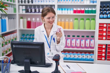 Foto de Joven mujer rubia farmacéutica usando la computadora sosteniendo pastillas sonriendo en la farmacia - Imagen libre de derechos