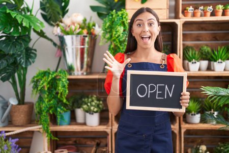 Photo for Young hispanic woman working at florist holding open sign celebrating victory with happy smile and winner expression with raised hands - Royalty Free Image