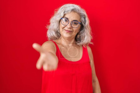 Foto de Mujer de mediana edad con el pelo gris de pie sobre fondo rojo sonriente alegre que ofrece la palma de la mano dando asistencia y aceptación. - Imagen libre de derechos