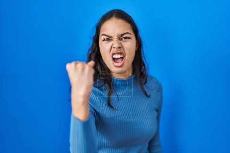 Photo for Young brazilian woman standing over blue isolated background angry and mad raising fist frustrated and furious while shouting with anger. rage and aggressive concept. - Royalty Free Image