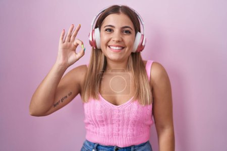 Téléchargez les photos : Jeune femme blonde écoutant de la musique à l'aide d'écouteurs souriant positif faire ok signe avec la main et les doigts. expression réussie. - en image libre de droit