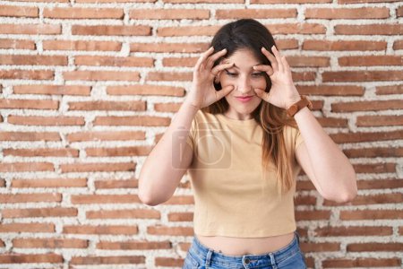Téléchargez les photos : Jeune femme brune debout sur un mur de briques essayant d'ouvrir les yeux avec les doigts, endormie et fatiguée pour la fatigue matinale - en image libre de droit