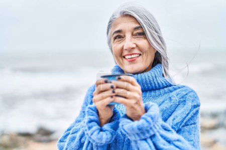Foto de Mujer de pelo gris de mediana edad sonriendo confiado bebiendo café en la playa - Imagen libre de derechos