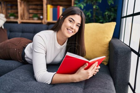Photo for Young beautiful hispanic woman reading book lying on sofa at home - Royalty Free Image
