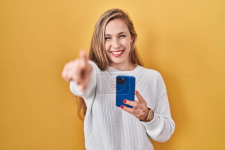 Téléchargez les photos : Jeune femme blonde en utilisant un message de dactylographie smartphone pointant vers vous et la caméra avec les doigts, souriant positif et joyeux - en image libre de droit