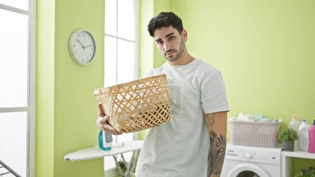 Foto de Joven hombre hispano sosteniendo canasta con ropa con expresión triste en la lavandería - Imagen libre de derechos