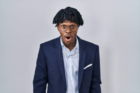 Téléchargez les photos : Jeune homme africain avec dreadlocks portant une veste d'affaires sur fond blanc effrayé et choqué par la surprise et l'expression étonnante, la peur et le visage excité. - en image libre de droit