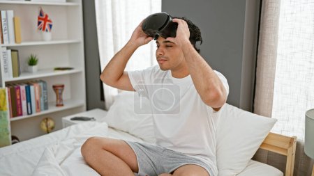 Foto de Hombre latino joven sentado en la cama con gafas de realidad virtual en el dormitorio - Imagen libre de derechos