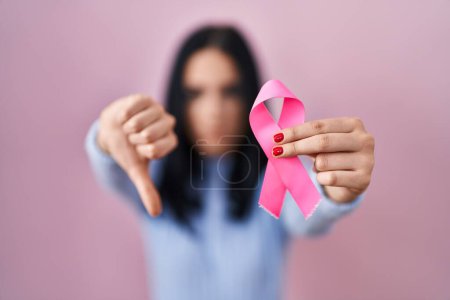 Photo for Hispanic woman holding pink cancer ribbon with angry face, negative sign showing dislike with thumbs down, rejection concept - Royalty Free Image