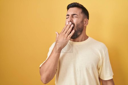 Photo for Handsome hispanic man standing over yellow background bored yawning tired covering mouth with hand. restless and sleepiness. - Royalty Free Image