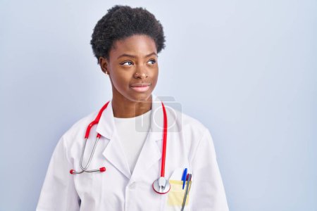 Téléchargez les photos : Femme afro-américaine portant l'uniforme de médecin et stéthoscope regardant de côté avec le sourire sur le visage, expression naturelle. riant confiant. - en image libre de droit