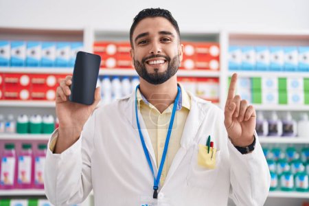 Foto de Joven hispano trabajando en farmacia mostrando la pantalla del teléfono inteligente sonriendo con una idea o pregunta señalando con el dedo con la cara feliz, número uno - Imagen libre de derechos