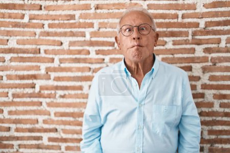 Téléchargez les photos : Homme âgé avec les cheveux gris debout sur le mur de briques faisant visage de poisson avec des lèvres, geste fou et comique. expression drôle. - en image libre de droit