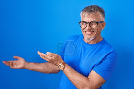 Foto de Hombre hispano con el pelo gris de pie sobre fondo azul invitando a entrar sonriente natural con la mano abierta - Imagen libre de derechos