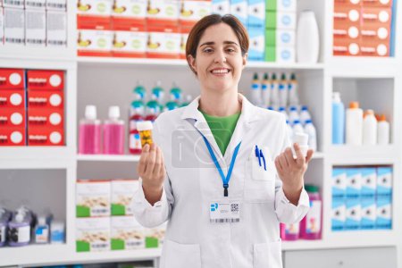 Téléchargez les photos : Jeune femme pharmacienne souriant confiant tenant des bouteilles de pilules à la pharmacie - en image libre de droit
