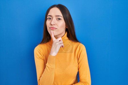 Téléchargez les photos : Jeune femme brune debout sur fond bleu pensant concentré sur le doute avec le doigt sur le menton et regardant vers le haut se demandant - en image libre de droit