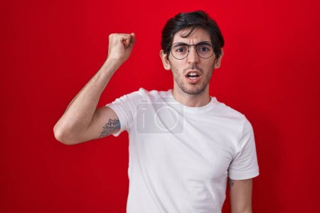 Téléchargez les photos : Jeune homme hispanique debout sur fond rouge en colère et fou levant le poing frustré et furieux tout en criant de colère. rage et concept agressif. - en image libre de droit