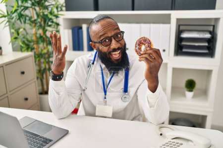 Photo for African american man working at dietitian clinic holding doughnut celebrating victory with happy smile and winner expression with raised hands - Royalty Free Image