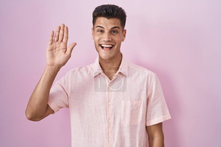 Foto de Joven hombre hispano de pie sobre fondo rosa renunciando a decir hola feliz y sonriente, gesto de bienvenida amistoso - Imagen libre de derechos