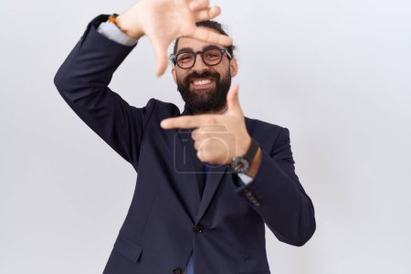Foto de Hombre hispano con barba vistiendo traje y corbata sonriente haciendo marco con manos y dedos con cara feliz. concepto de creatividad y fotografía. - Imagen libre de derechos