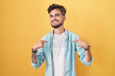 Foto de Joven hombre hispano con tatuajes de pie sobre fondo amarillo mirando confiado con sonrisa en la cara, señalándose con los dedos orgullosos y felices. - Imagen libre de derechos