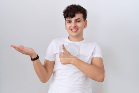 Téléchargez les photos : Jeune homme non binaire portant un t-shirt blanc décontracté montrant la main de la paume et faisant un geste correct avec les pouces levés, souriant heureux et joyeux - en image libre de droit
