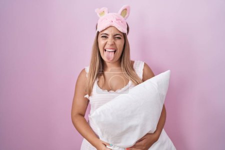 Foto de Mujer rubia joven con pijama abrazando almohada sacando la lengua feliz con expresión divertida. concepto de emoción. - Imagen libre de derechos