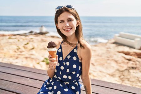 Téléchargez les photos : Jeune femme blonde touriste tenant crème glacée assis sur le banc au bord de la mer - en image libre de droit