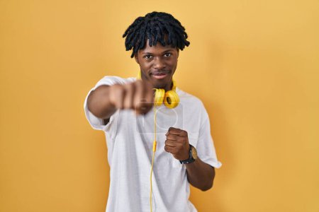 Téléchargez les photos : Jeune homme africain avec dreadlocks debout sur fond jaune coup de poing poing pour se battre, attaque agressive et en colère, menace et violence - en image libre de droit