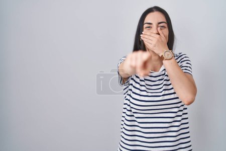 Photo for Young brunette woman wearing striped t shirt laughing at you, pointing finger to the camera with hand over mouth, shame expression - Royalty Free Image