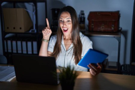 Téléchargez les photos : Jeune femme brune travaillant au bureau la nuit pointant du doigt avec une idée réussie. Sorti et heureux. numéro un. - en image libre de droit