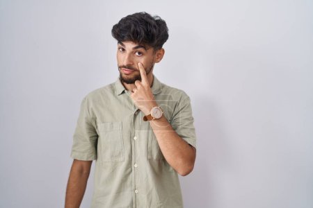 Foto de Hombre árabe con barba de pie sobre fondo blanco apuntando al ojo observándote gesto, expresión sospechosa - Imagen libre de derechos